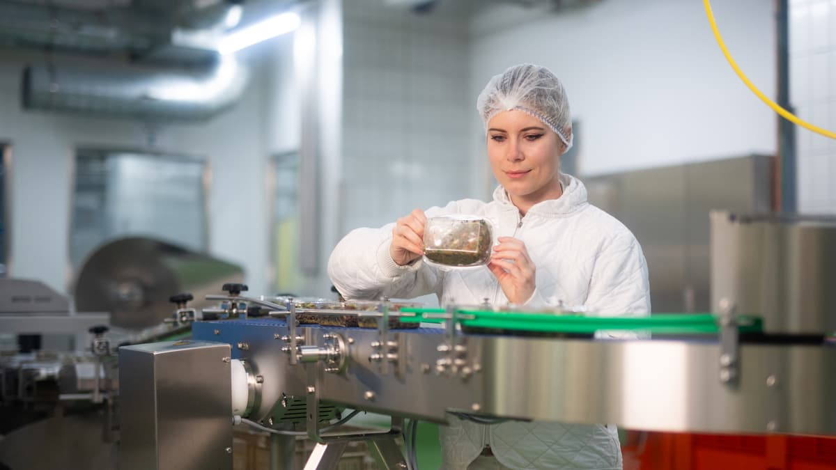 Frau mit Haube steht an Produktionsanlage und hält Verpackung mit Essen in der Hand