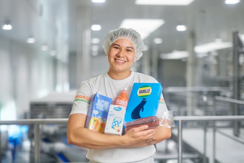 Mitarbeitende hält diverse Kult-Migros-Produkte in den Armen und lächelt in Kamera
