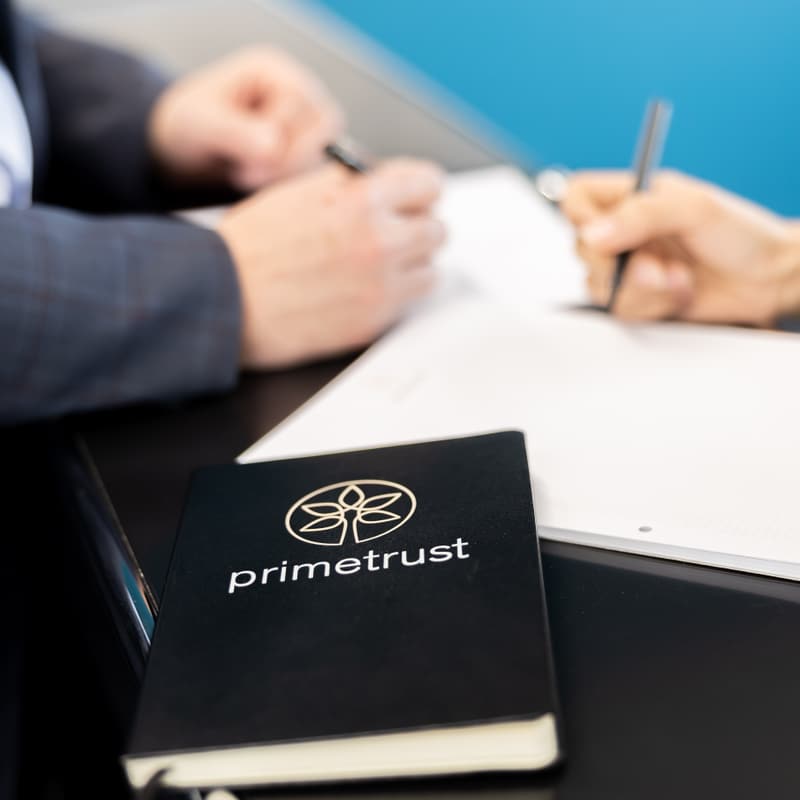 Zwei Personen schreiben etwas in Notizheft, vor ihnen liegt ein Notizbuch mit der Aufschrift «primetrust»