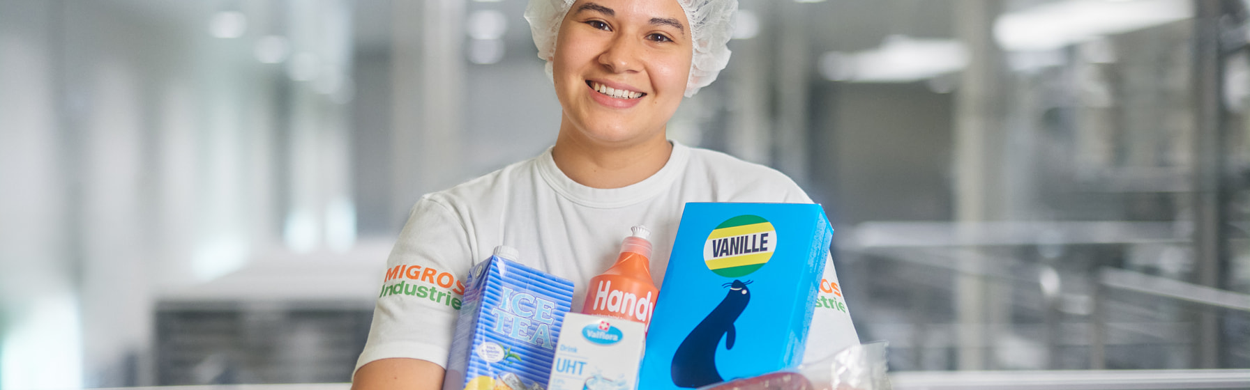 Mitarbeitende hält diverse Kult-Migros-Produkte in den Armen und lächelt in Kamera